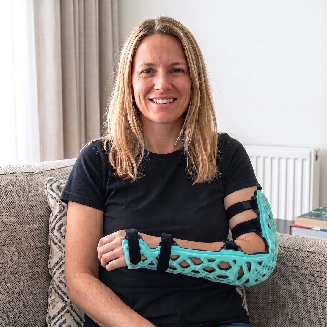 The same woman on the same sofa wearing a teal FlexiSplint lattice forearm shell, no sling, smiling