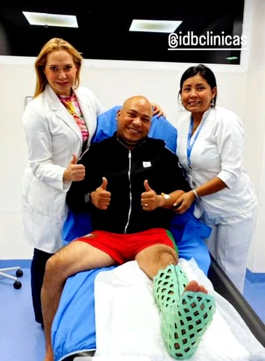A seated patient giving a thumbs up with two clinicians beside him, wearing a green FlexiOH short leg immobilizer